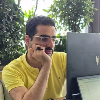 Mahdi in a yellow polo and clear glasses, chin on his hand, looking at a laptop.